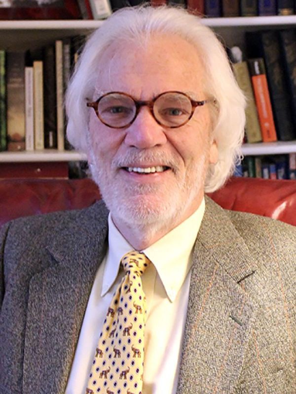 A portrait of a man with white hair in front of a bookshelf 