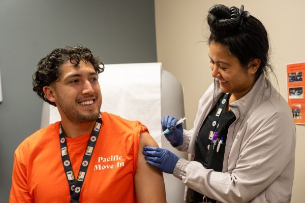 Man in an orange "Pacific Move-In" shirt receiving an immunization from a Nurse Practicioner