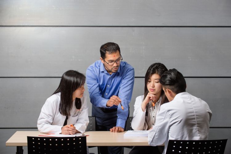 a professor stands next to a table with three seated students