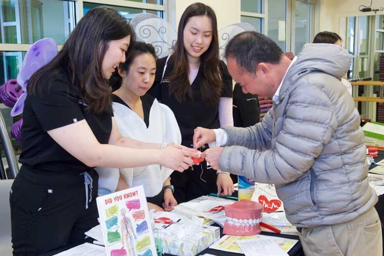 Three women dental students wearing scrubs holding a model of teeth for a man to practice flossing