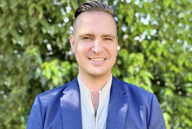 A man in a suit smiles in front of a wall of greenery