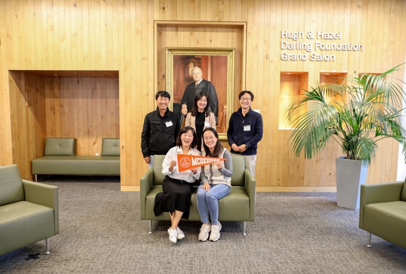 Five Korean individuals pose for a photo. Three are standing, and two are sitting and holding an orange pennant. 