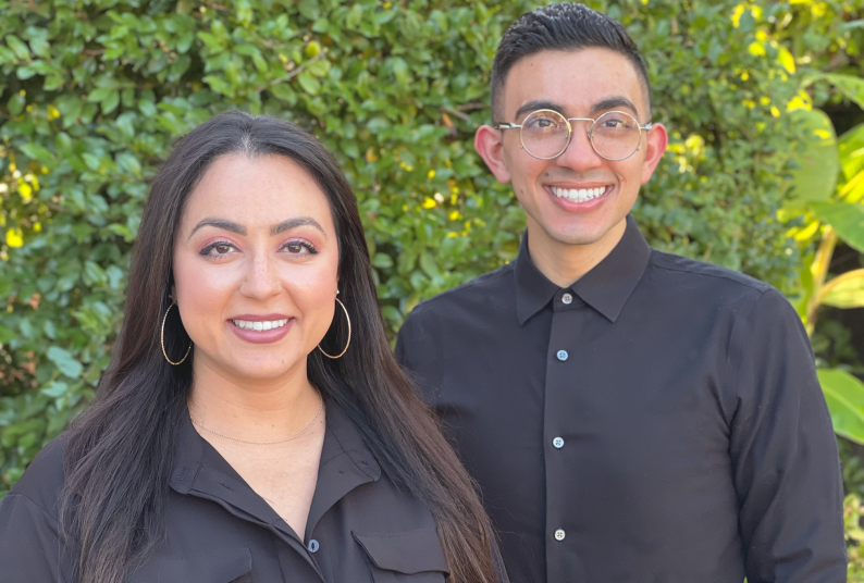 Two individuals wearing black pose for a photo in front of shrubbery 