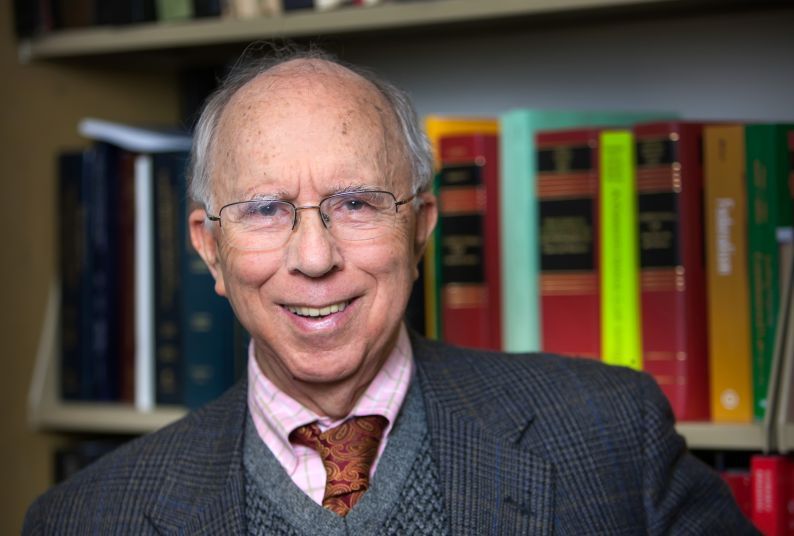 An older gentleman in a suit poses in front of a bookcase.