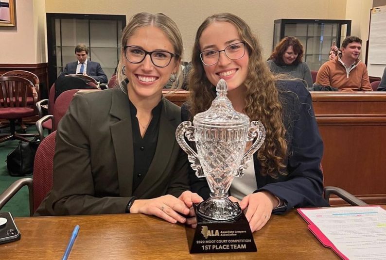 Two female students hold a trophy in a courtroom while seated