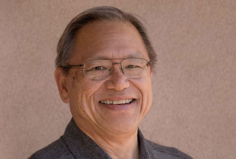 A portrait of a man wearing a gray polo shirt in front of a tan neutral background 