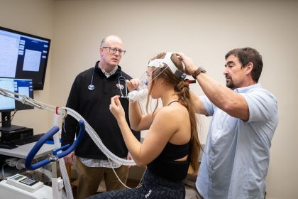 two professors stand next to a student hooked up to a machine for exercise testing