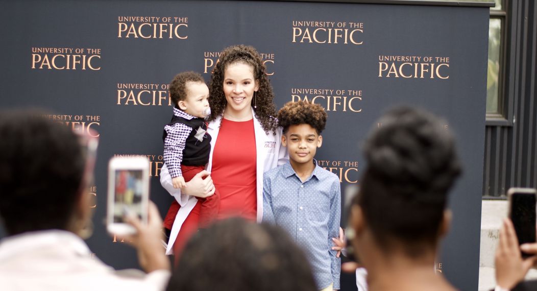 Photo shows a smiling PA student holding her young son while her older son stands beside her as people in the audience snap their photo.