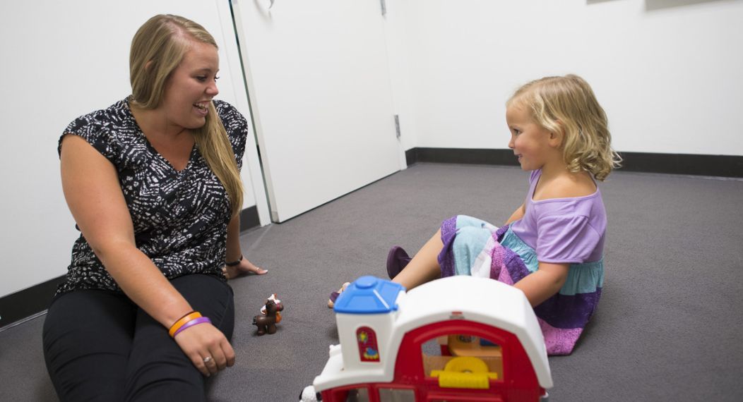 The image shows a student sitting on the floor and talking to a child while playing with toys.